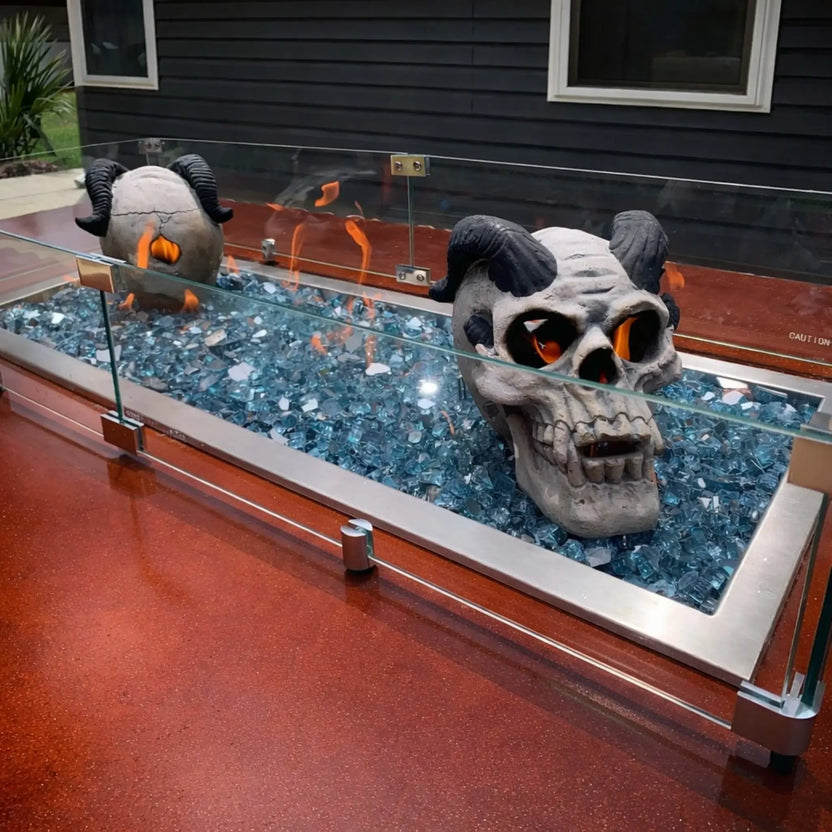 Linear fire pit installation filled with blue fire glass and decorative skull accents, enclosed by a tempered glass wind guard, creating a bold custom fire feature set into a red concrete patio surface.