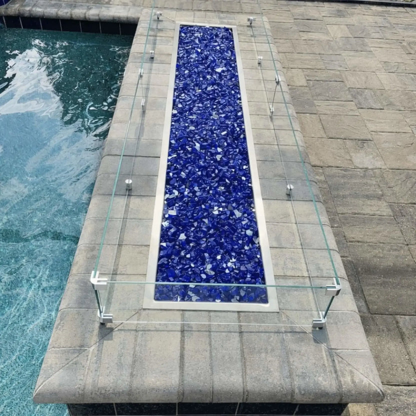 Top-down view of a narrow rectangular fire pit near a swimming pool, featuring deep blue fire glass and a clear tempered glass wind guard mounted on stainless steel brackets along a stone pool ledge.