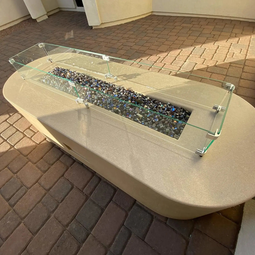 Outdoor linear fire pit table with a clear tempered glass wind guard installed, filled with reflective fire glass and set into a smooth concrete-style patio table on paver flooring.