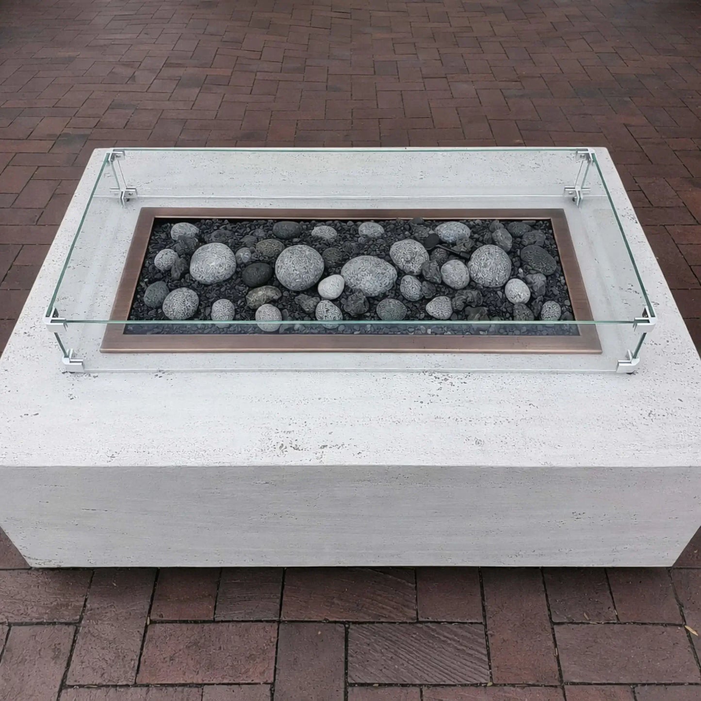 Minimalist white concrete fire pit table with a rectangular burner filled with smooth gray lava stones, surrounded by a low-profile tempered glass wind guard on a brick patio.