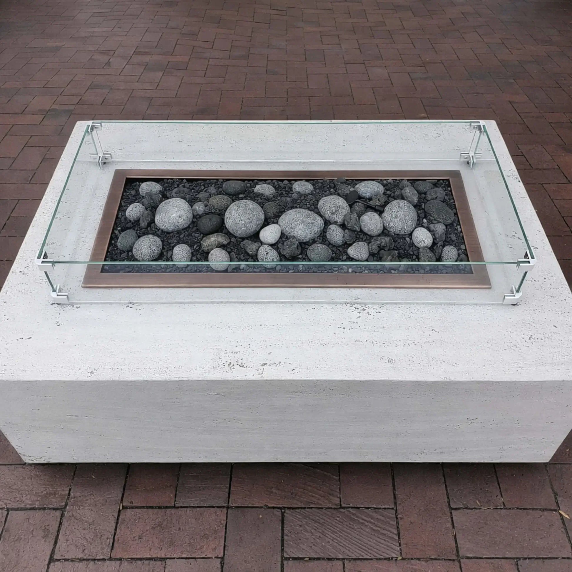 Minimalist white concrete fire pit table with a rectangular burner filled with smooth gray lava stones, surrounded by a low-profile tempered glass wind guard on a brick patio.