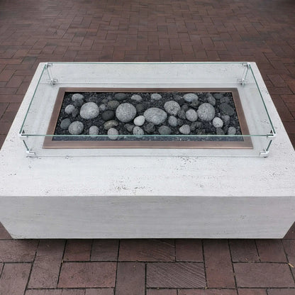 Minimalist white concrete fire pit table with a rectangular burner filled with smooth gray lava stones, surrounded by a low-profile tempered glass wind guard on a brick patio.