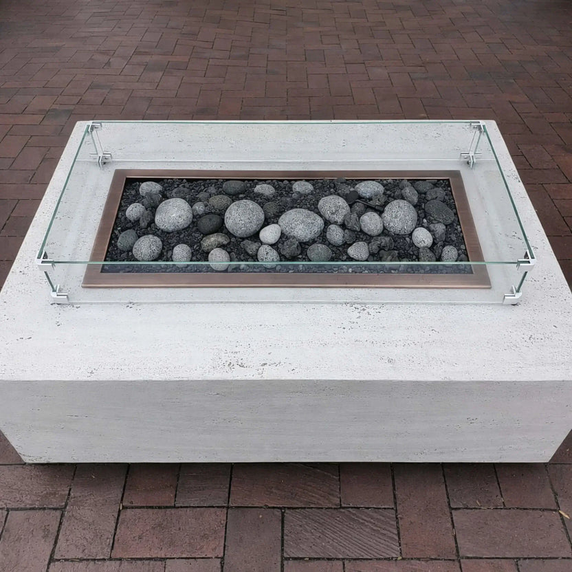 Minimalist white concrete fire pit table with a rectangular burner filled with smooth gray lava stones, surrounded by a low-profile tempered glass wind guard on a brick patio.