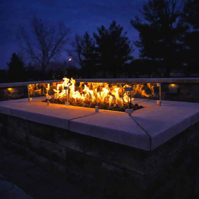 Outdoor stone fire pit at dusk featuring bright yellow flames rising above fire media, fully enclosed by a clear glass wind guard, set against a dark sky and landscaped backyard setting.