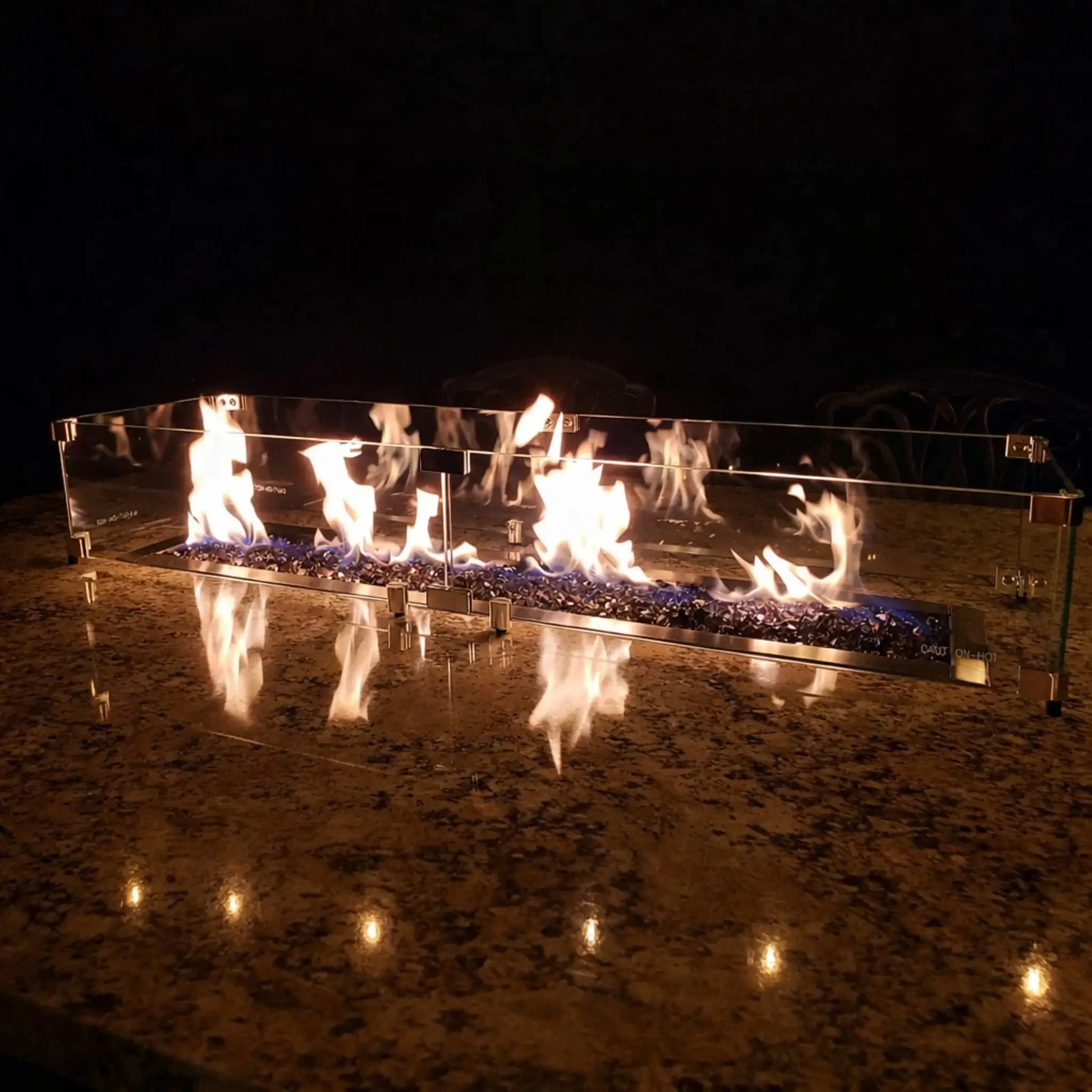 Linear fire pit with blue fire glass and lively flames reflected on a polished stone surface, protected by a tempered glass wind guard, creating a dramatic nighttime fire feature on an outdoor countertop.