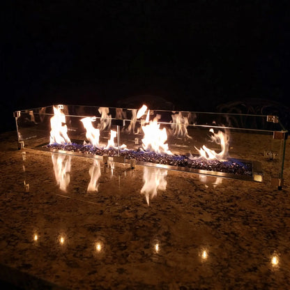 Linear fire pit with blue fire glass and lively flames reflected on a polished stone surface, protected by a tempered glass wind guard, creating a dramatic nighttime fire feature on an outdoor countertop.
