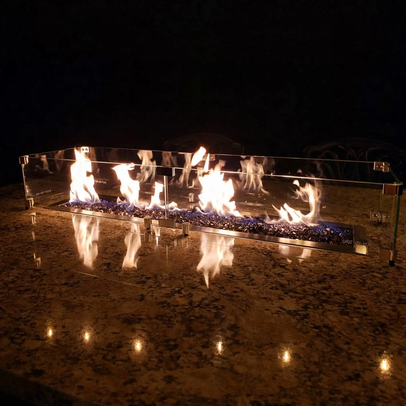 Linear fire pit with blue fire glass and lively flames reflected on a polished stone surface, protected by a tempered glass wind guard, creating a dramatic nighttime fire feature on an outdoor countertop.