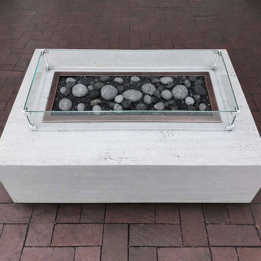 Modern rectangular fire pit table finished in white concrete with a linear stainless steel burner tray filled with smooth gray river rocks, protected by a clear tempered glass wind guard, set on a brick patio for a clean contemporary outdoor look.