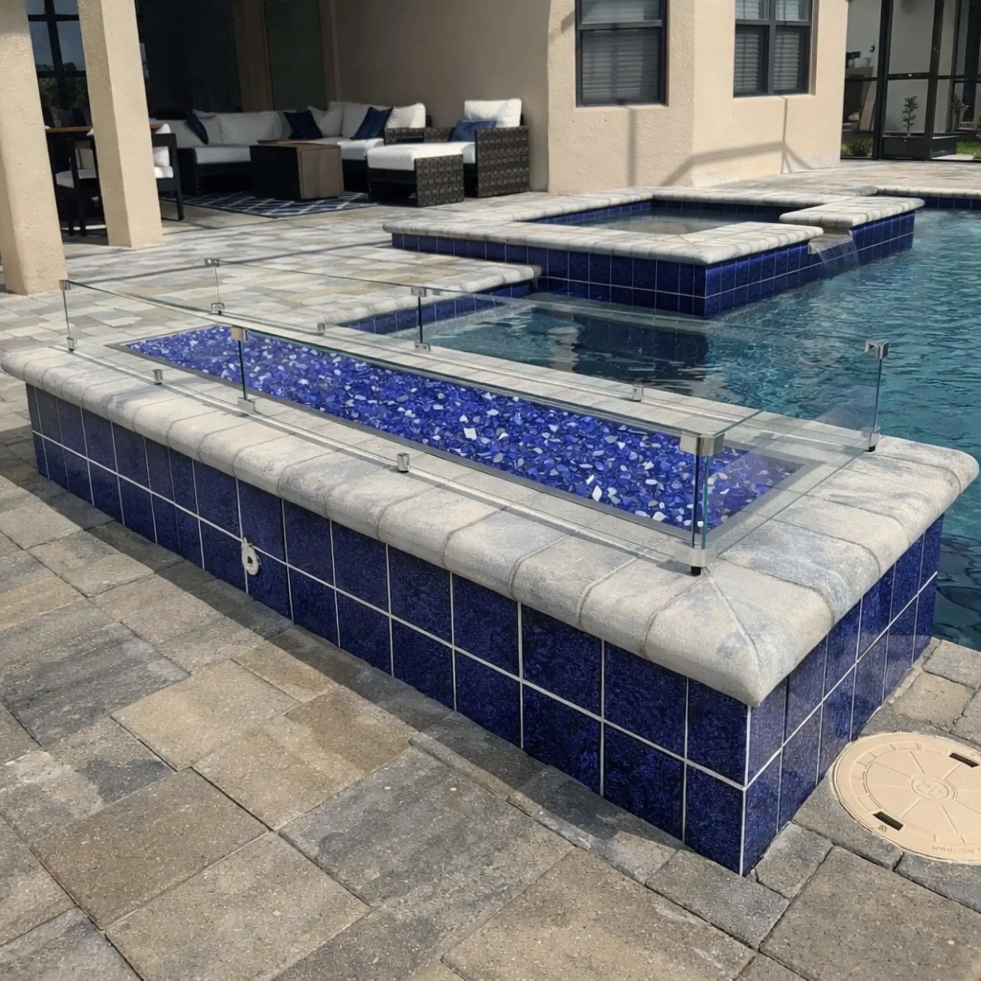 Linear fire pit installed along a pool edge with blue fire glass media, stainless steel burner pan, and clear glass wind guard, integrated into a tiled pool surround with water and patio seating visible in the background.
