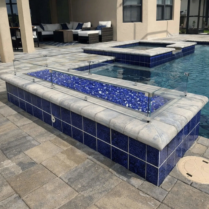 Linear fire pit installed along a pool edge with blue fire glass media, stainless steel burner pan, and clear glass wind guard, integrated into a tiled pool surround with water and patio seating visible in the background.
