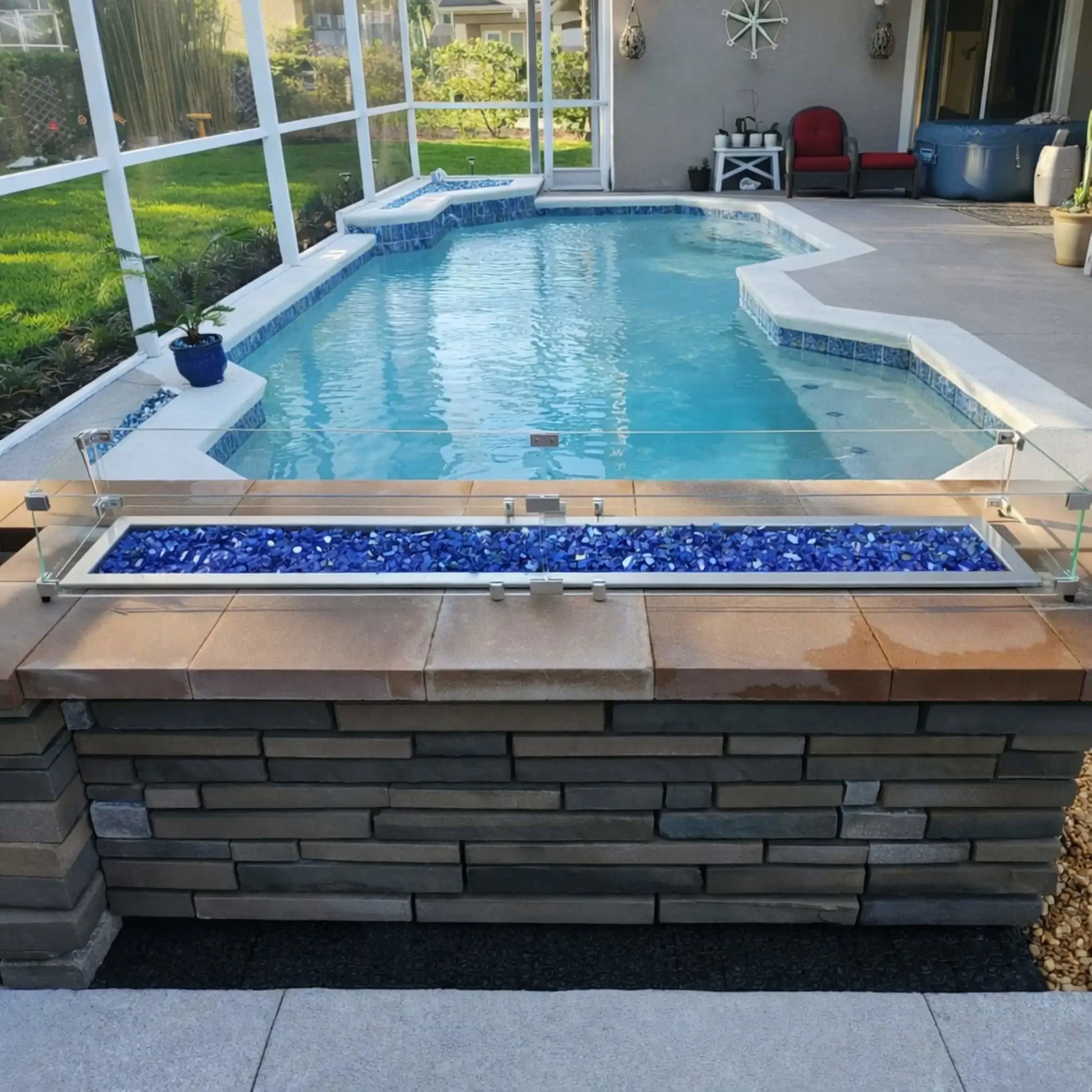 Backyard pool area featuring a long rectangular fire pit with blue glass fire media, stainless steel burner, and tempered glass wind guard mounted on a stone structure overlooking a screened-in swimming pool.