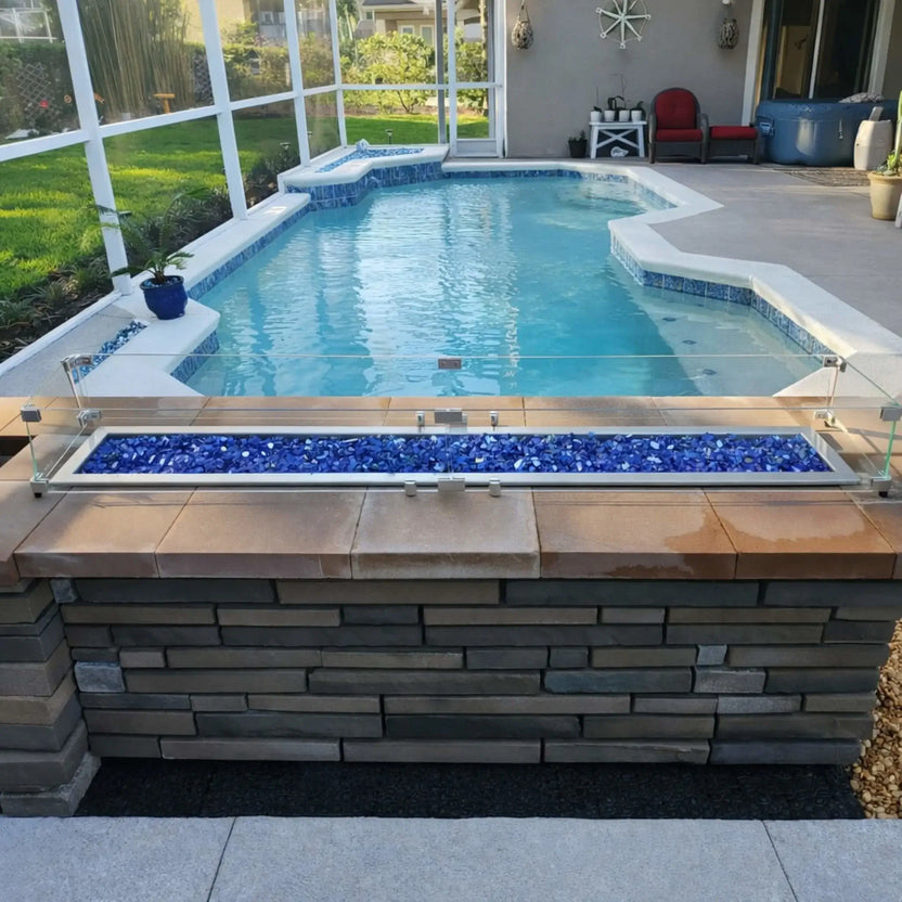 Backyard pool area featuring a long rectangular fire pit with blue glass fire media, stainless steel burner, and tempered glass wind guard mounted on a stone structure overlooking a screened-in swimming pool.