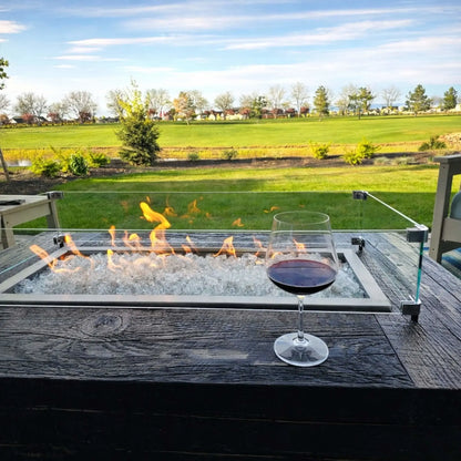 Outdoor linear fire pit table with clear fire glass and a glass wind guard, showcasing active flames beside a wine glass on a dark wood surface, overlooking an open green landscape for an elegant patio entertaining setting.