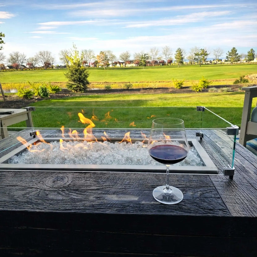 Outdoor linear fire pit table with clear fire glass and a glass wind guard, showcasing active flames beside a wine glass on a dark wood surface, overlooking an open green landscape for an elegant patio entertaining setting.