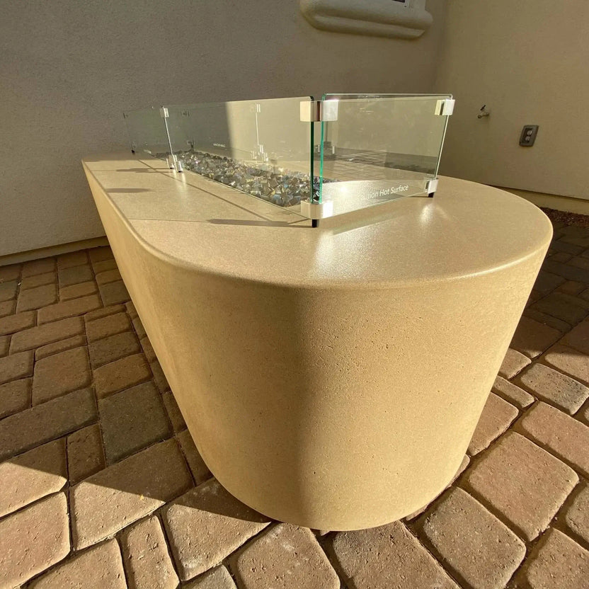 Side-angle view of a linear fire pit featuring a tempered glass wind guard mounted on a curved concrete fire table, showcasing reflective fire glass in a sunlit outdoor patio.