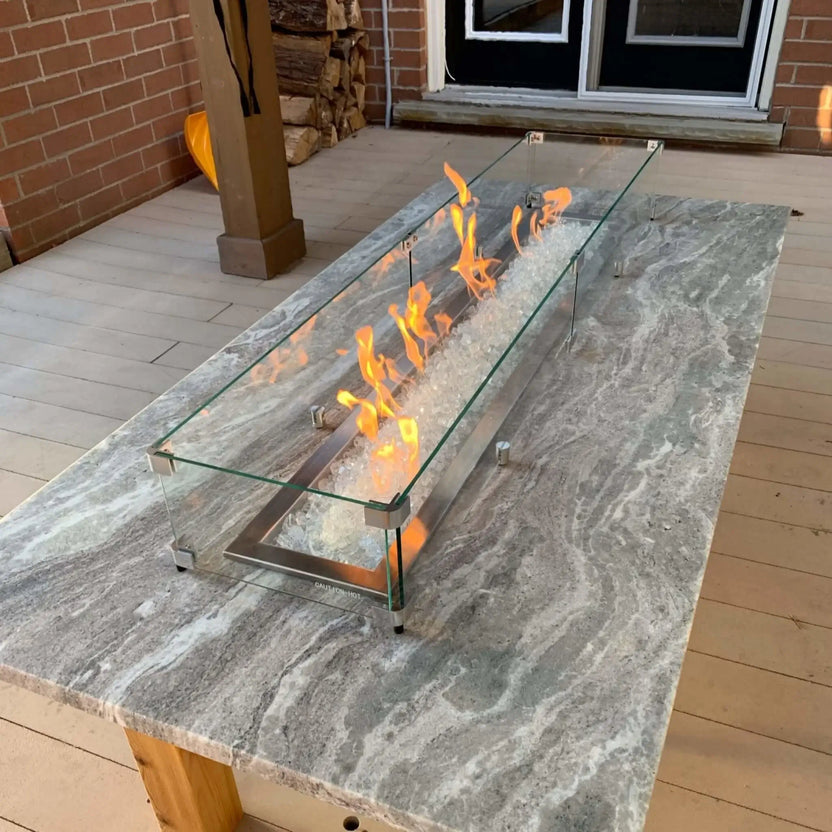Long rectangular fire pit with clear tempered glass wind guard and steady flames, set into a stone countertop with a stainless steel burner pan and white fire glass on a covered patio.