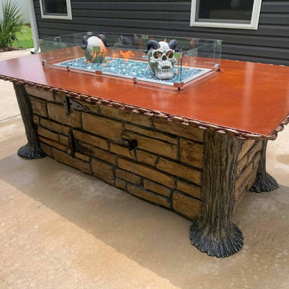 Decorative linear fire pit table with a tempered glass wind guard, blue fire glass, and sculptural accents inside the burner, mounted on a custom stone base in an outdoor living space.