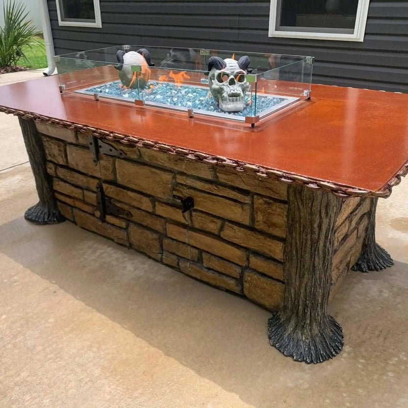 Decorative linear fire pit table with a tempered glass wind guard, blue fire glass, and sculptural accents inside the burner, mounted on a custom stone base in an outdoor living space.