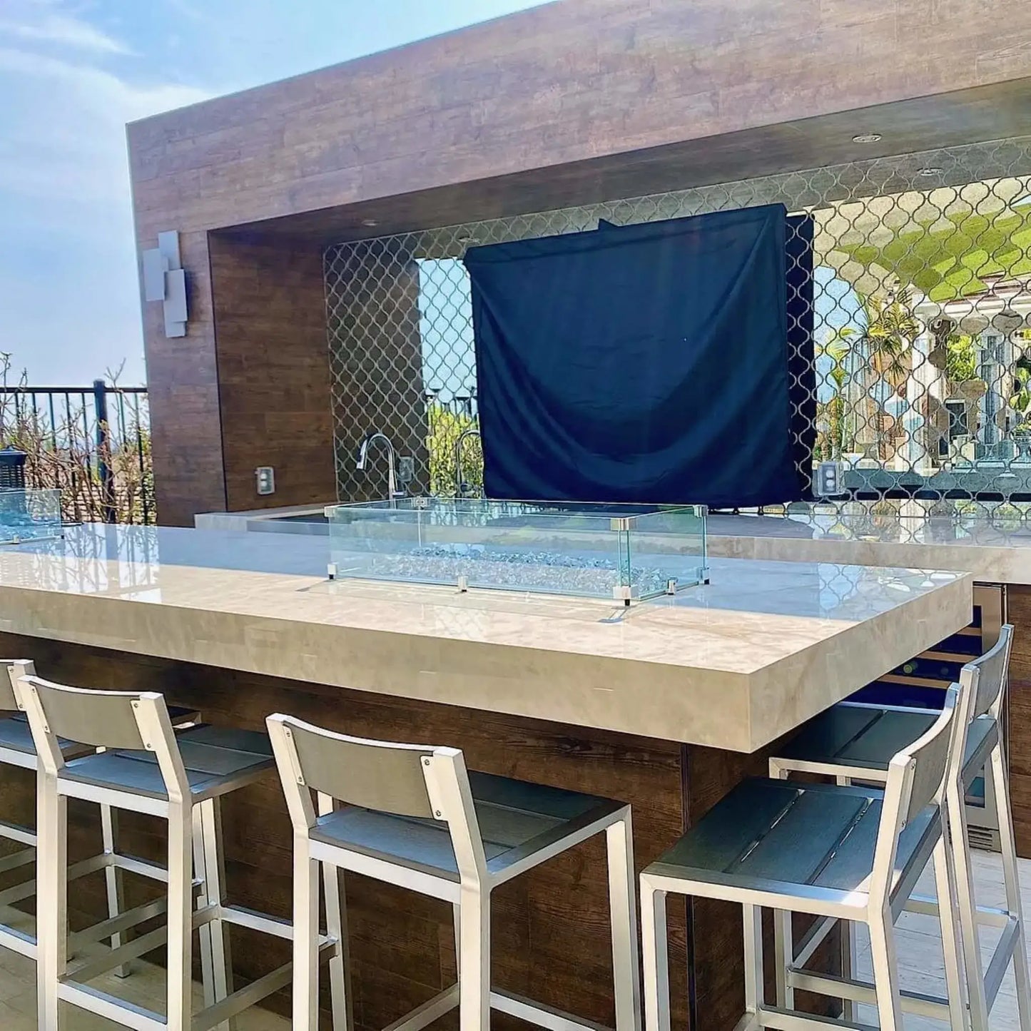 Modern outdoor kitchen bar with a linear rectangular fire pit featuring a clear tempered glass wind guard, set into a light stone countertop with bar seating, creating a sleek and contemporary outdoor entertaining space.
