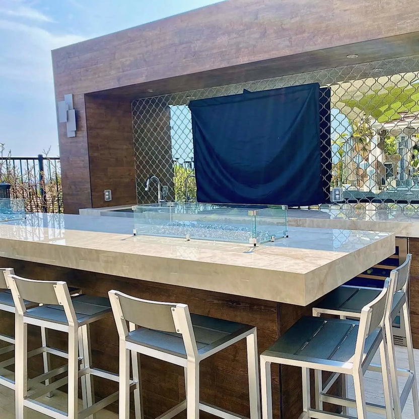 Modern outdoor kitchen bar with a linear rectangular fire pit featuring a clear tempered glass wind guard, set into a light stone countertop with bar seating, creating a sleek and contemporary outdoor entertaining space.