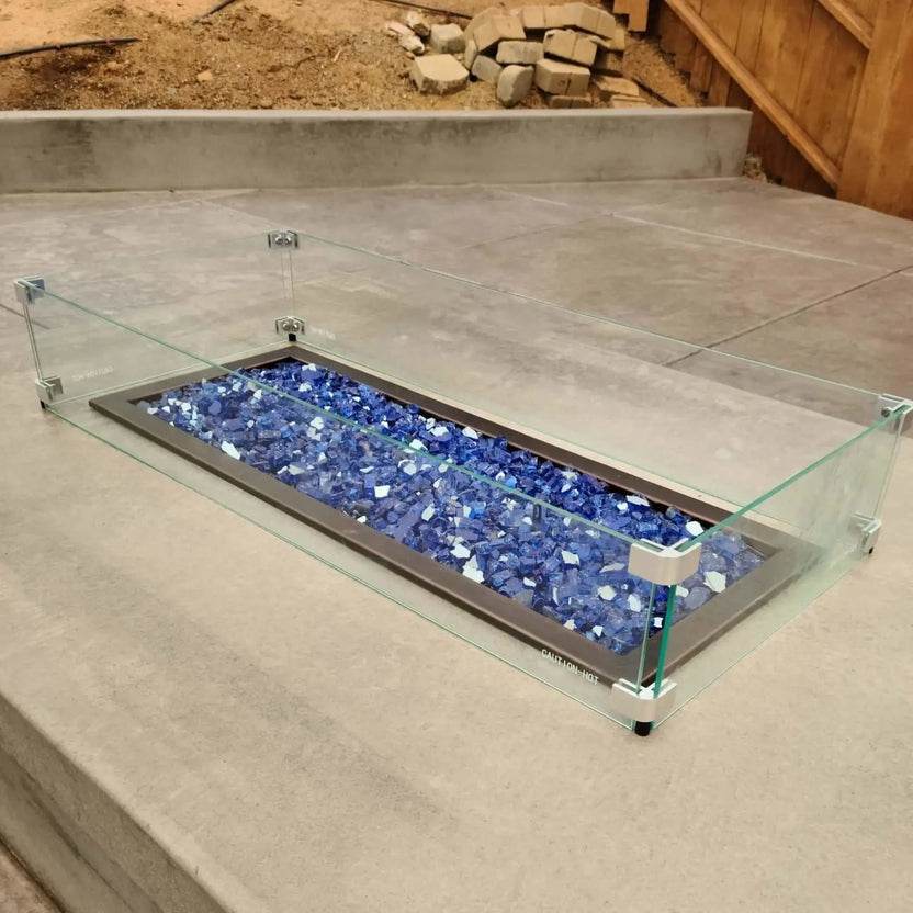 Close-up view of a linear rectangular fire pit with blue fire glass and a clear tempered glass wind guard, installed in a concrete patio setting, highlighting clean lines and modern outdoor fire feature design.