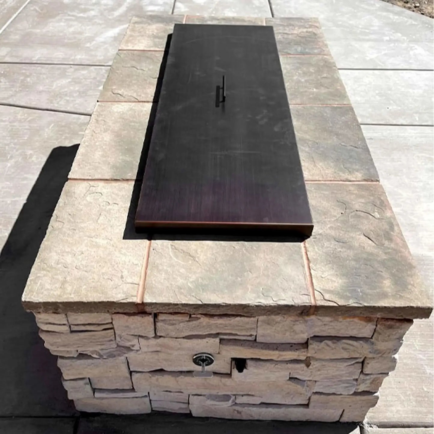 Oil rubbed bronze rectangular fire pit burner cover installed in a stone paver fire feature, shown with spark ignition hardware beneath for a clean, finished outdoor look.