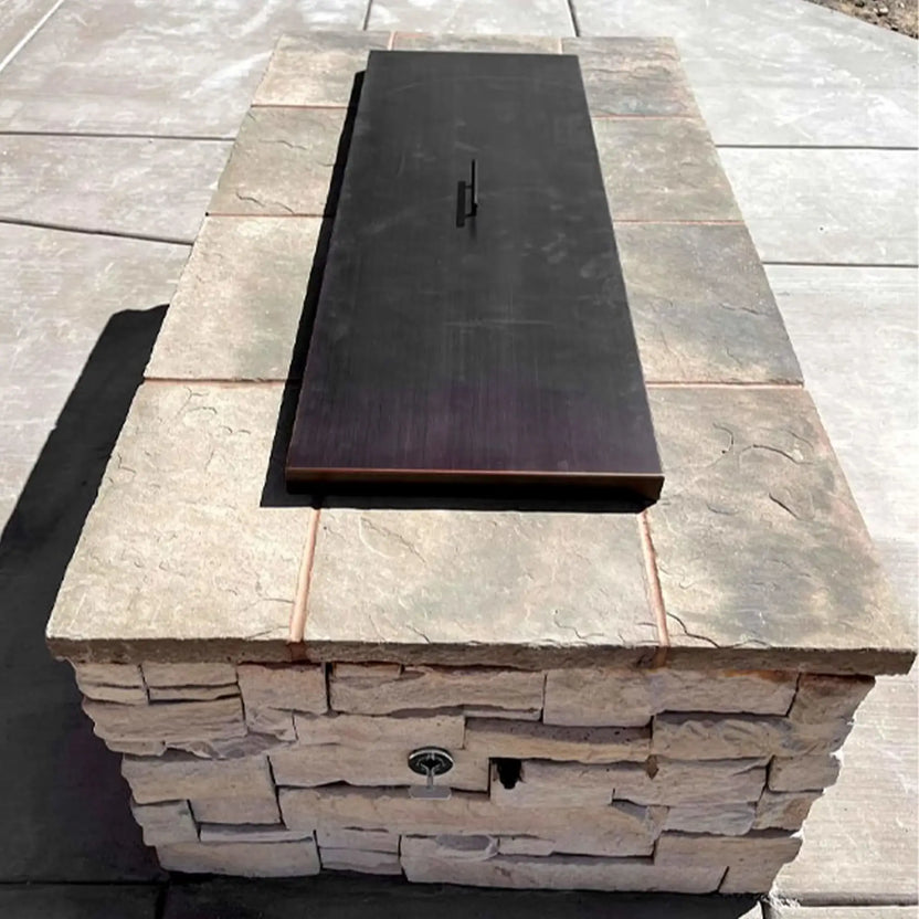Oil rubbed bronze rectangular fire pit burner cover installed in a stone paver fire feature, shown with spark ignition hardware beneath for a clean, finished outdoor look.