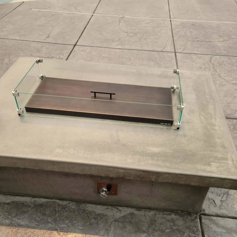 Linear oil rubbed bronze burner cover paired with a clear glass wind guard, installed on a stone fire pit with spark ignition components concealed below the cover.