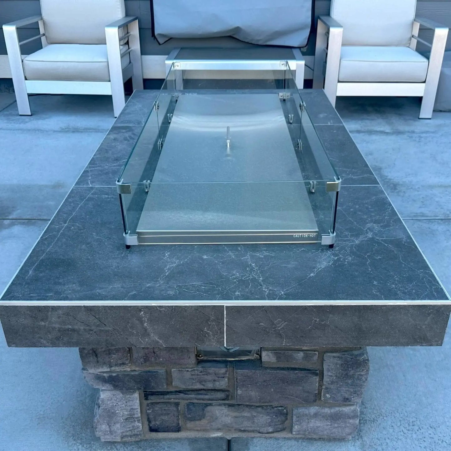 Rectangular stainless steel linear fire pit burner cover installed within a modern paver fire pit, featuring a clear glass wind guard secured at each corner, designed to protect the burner while creating a clean, finished tabletop surface.