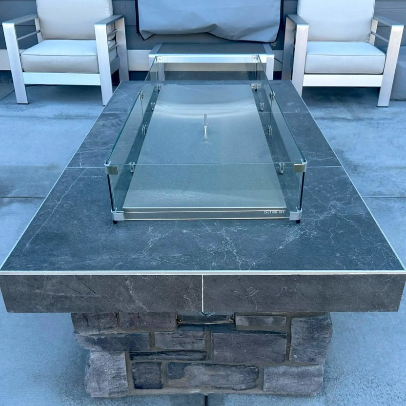 Rectangular stainless steel linear fire pit burner cover installed within a modern paver fire pit, featuring a clear glass wind guard secured at each corner, designed to protect the burner while creating a clean, finished tabletop surface.