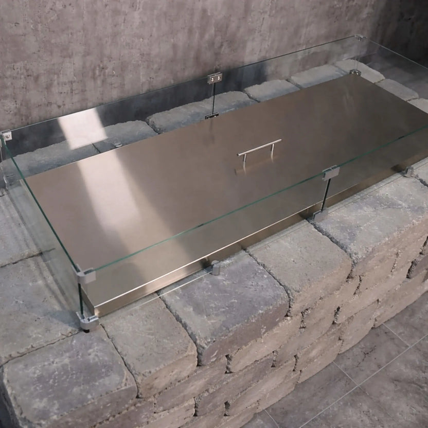 Rectangular stainless steel linear fire pit burner cover installed within a modern paver fire pit, featuring a clear glass wind guard secured at each corner, designed to protect the burner while creating a clean, finished tabletop surface.