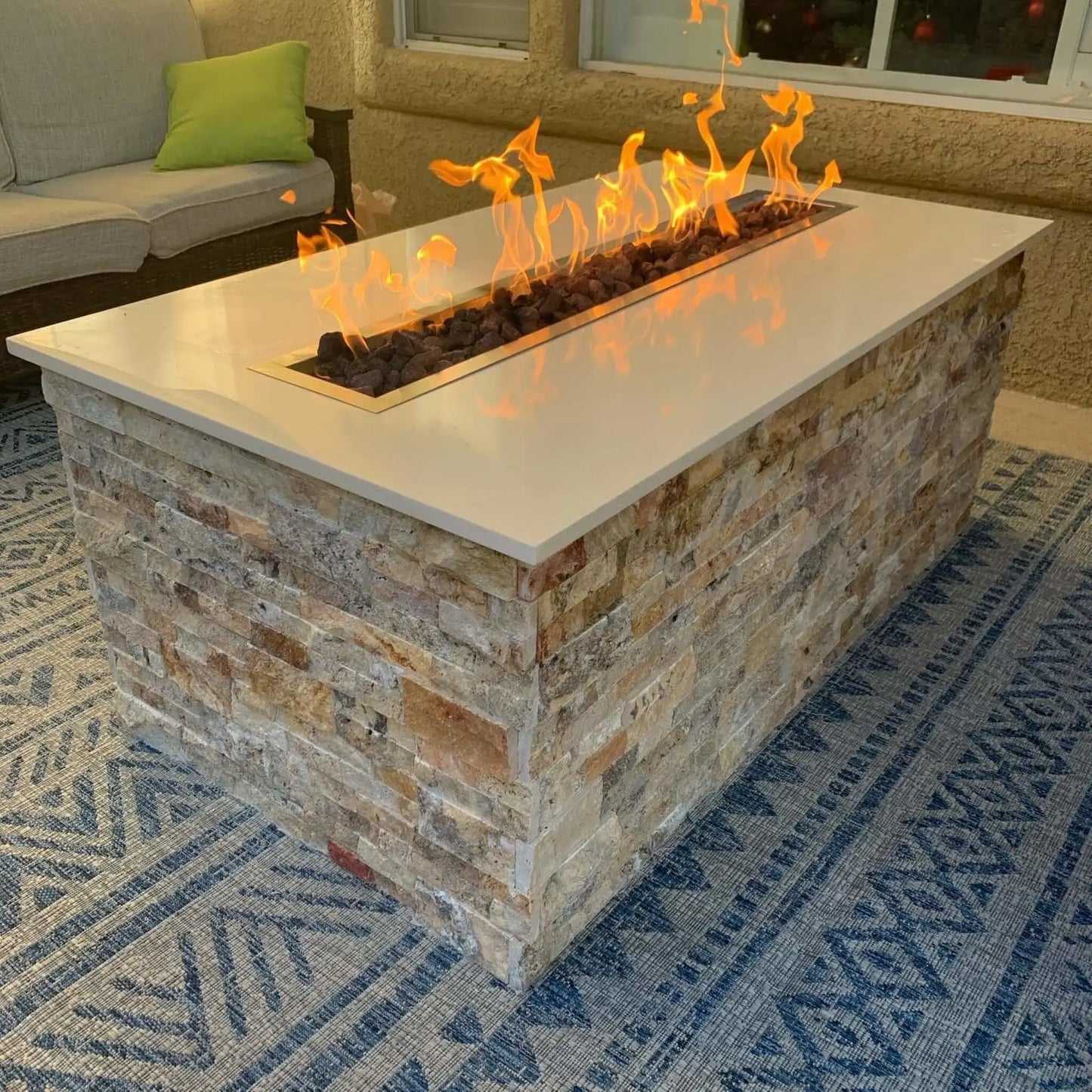 Rectangular outdoor fire pit table featuring a stainless steel linear burner pan installed flush into a smooth stone countertop, producing tall, even flames over lava rock in a modern patio seating area.