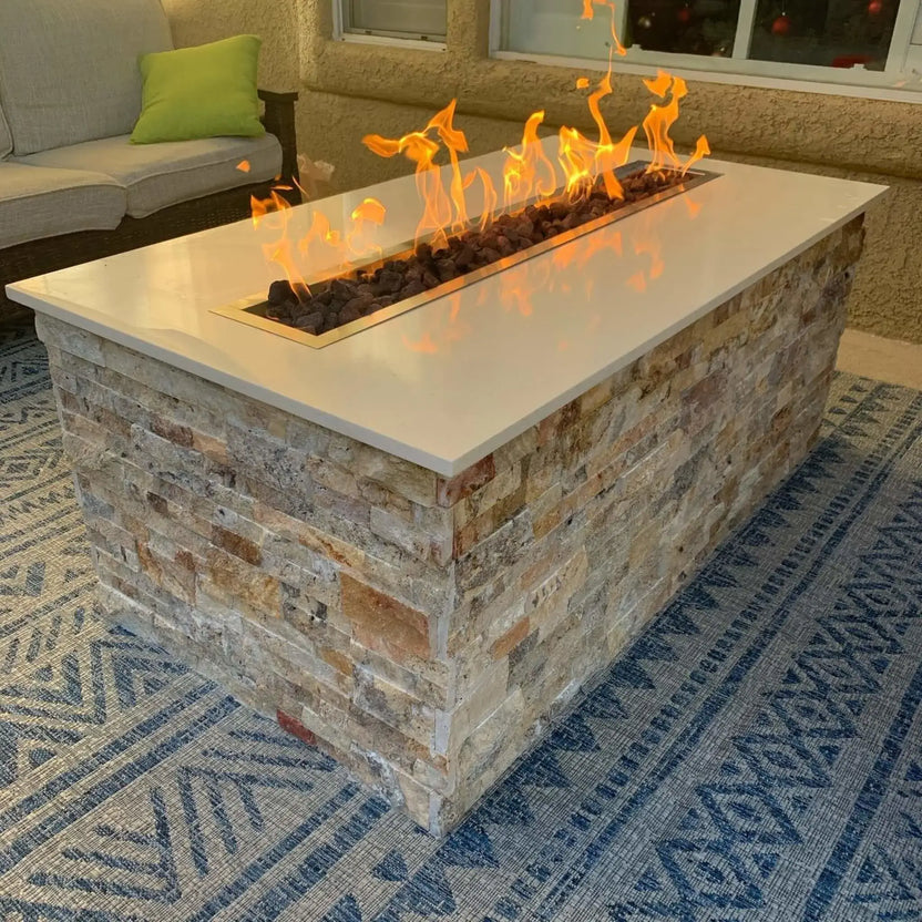 Rectangular outdoor fire pit table featuring a stainless steel linear burner pan installed flush into a smooth stone countertop, producing tall, even flames over lava rock in a modern patio seating area.