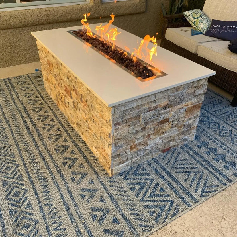 Modern outdoor fire pit with a long stainless steel linear burner pan set into a stacked stone base, showcasing vibrant flames running the full length of the fire feature on a covered patio.