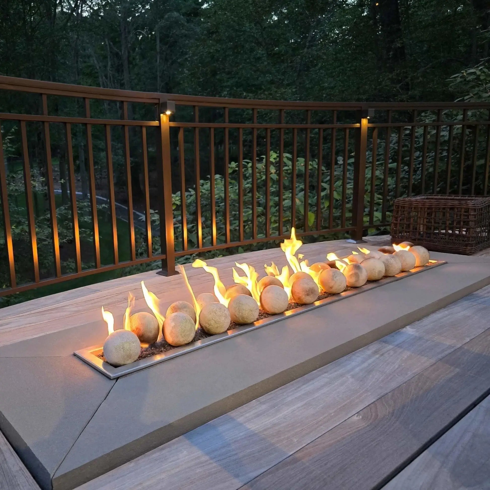 Custom-built outdoor fire pit table with a stainless steel linear burner pan centered in a stone enclosure, surrounded by patio furniture and decorative pavers in a landscaped backyard setting.