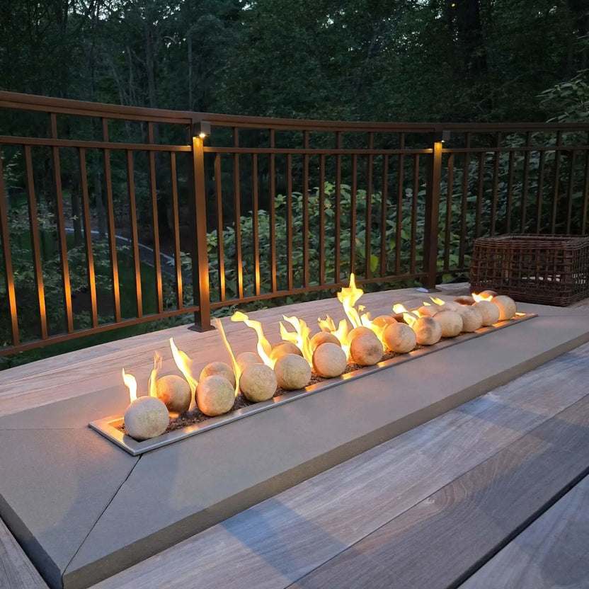 Custom-built outdoor fire pit table with a stainless steel linear burner pan centered in a stone enclosure, surrounded by patio furniture and decorative pavers in a landscaped backyard setting.