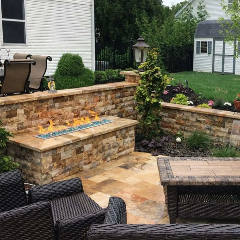 Contemporary outdoor fire feature featuring a stainless steel linear burner pan filled with smooth fire stones, installed in a concrete fire pit table on a deck overlooking a wooded landscape at dusk.