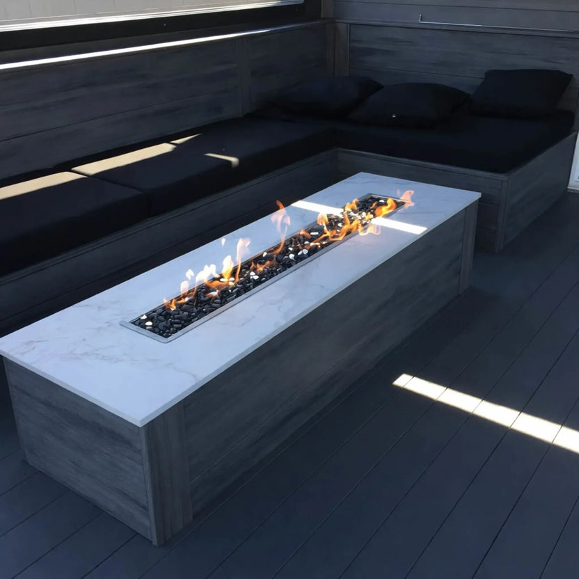 Backyard fire pit built with natural stone walls and a stainless steel linear burner pan, producing a clean ribbon of flame set against a landscaped garden and outdoor seating area.