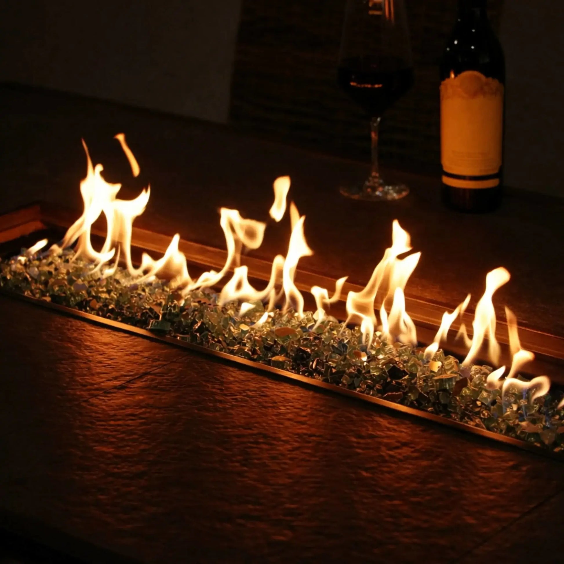 Indoor or covered outdoor linear fire feature with a stainless steel burner pan filled with fire glass, creating a continuous flame display along a wood surface beside wine glasses for an upscale, modern ambiance.