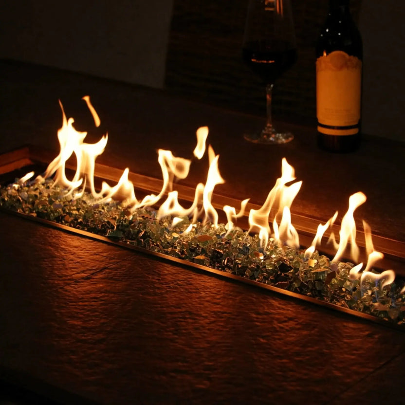 Indoor or covered outdoor linear fire feature with a stainless steel burner pan filled with fire glass, creating a continuous flame display along a wood surface beside wine glasses for an upscale, modern ambiance.