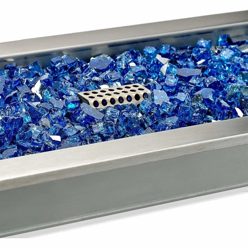 Close-up of the ignitor cage inside a stainless steel linear fire pit pan filled with blue cobalt fire glass, showing the protected ignition component surrounded by reflective tempered glass media.