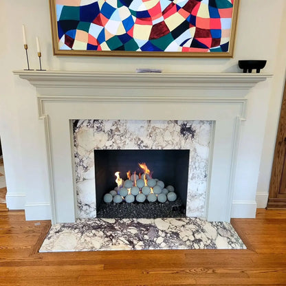 Modern indoor fireplace with a white mantel and marble surround, featuring Cape Grey fire balls stacked over black lava rock with flames rising between the spheres.