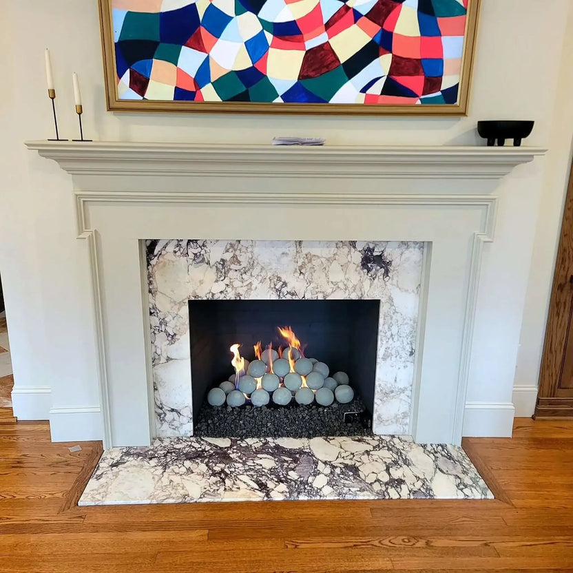 Modern indoor fireplace with a white mantel and marble surround, featuring Cape Grey fire balls stacked over black lava rock with flames rising between the spheres.