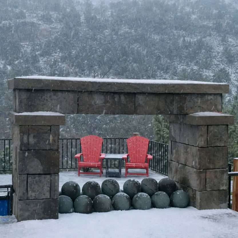 Outdoor stone fireplace in falling snow with dark fire balls lined along the base, framed by a snowy patio scene with two red Adirondack chairs in the background.