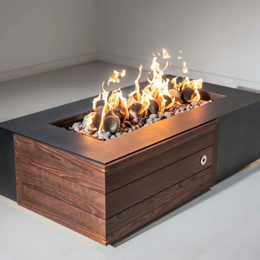 Wider patio view of a sleek fire pit table in front of a light-colored sectional sofa, displaying Cape Grey and darker fire balls resting in the burner with no flames.