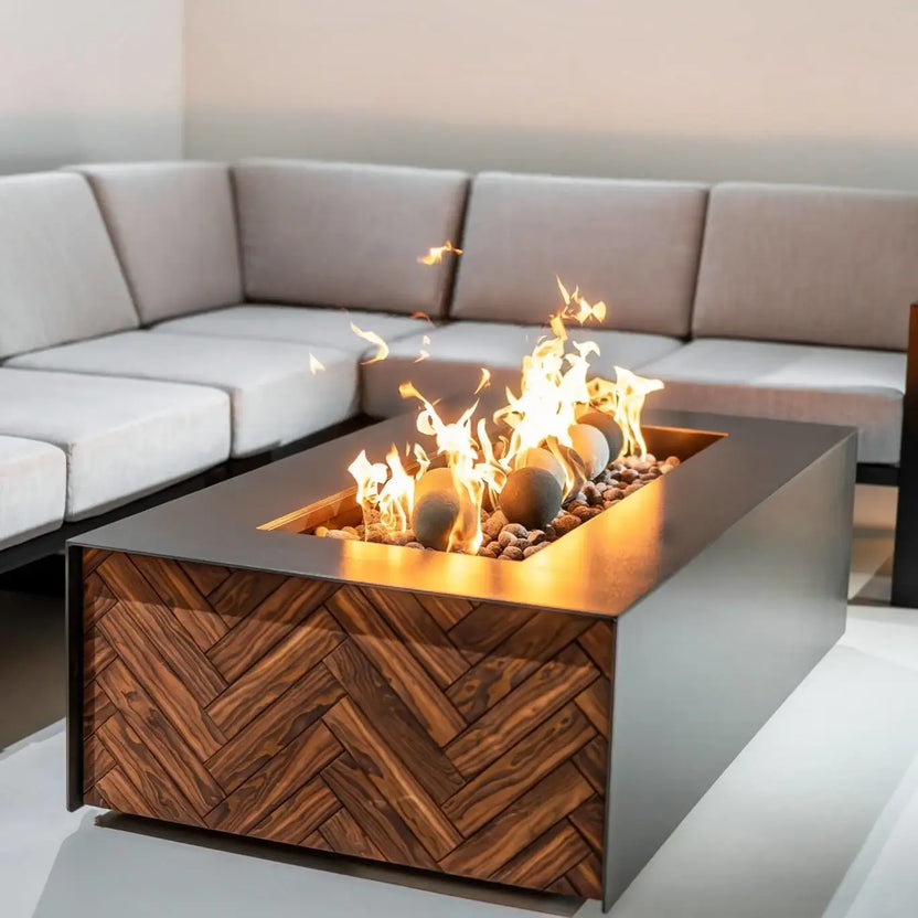 Contemporary rectangular fire pit table with a wood-pattern panel and dark metal surround, flames rising over round fire balls and small lava rock inside the burner.