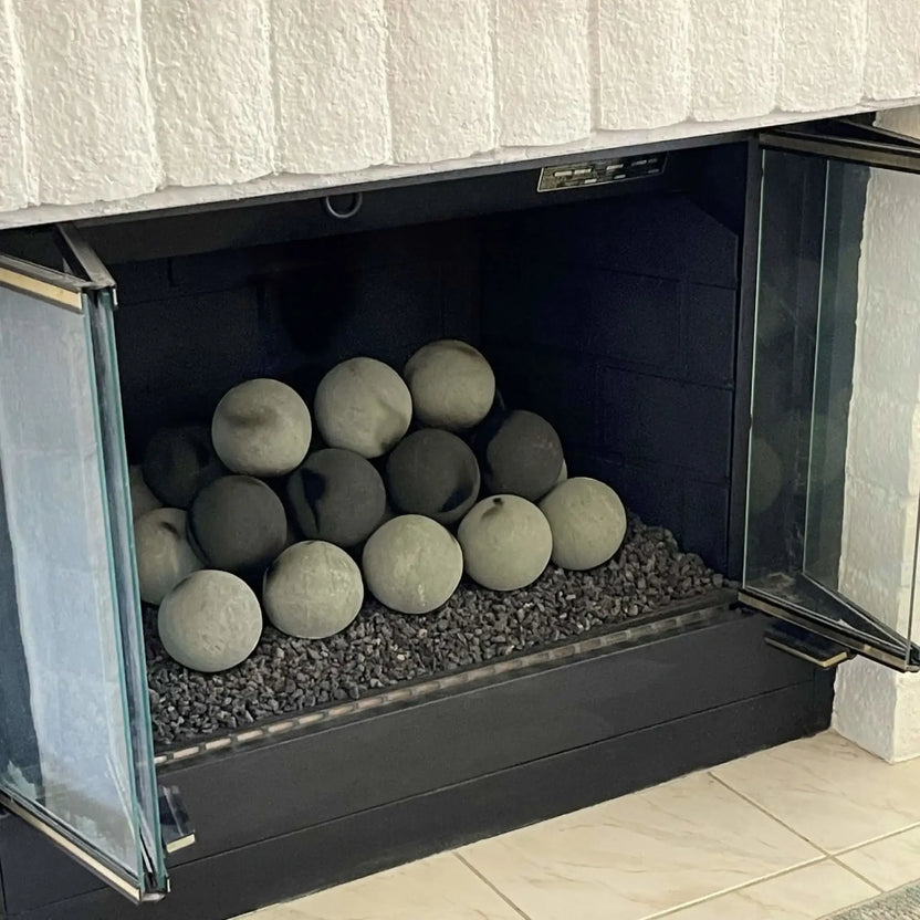 Glass-front indoor fireplace filled with round Cape Grey and darker fire balls on black lava rock, shown with the doors open and the burner media neatly arranged.