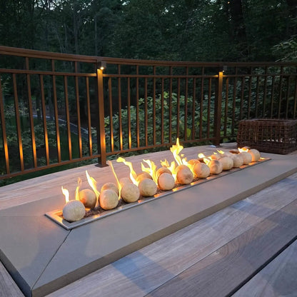 Linear gas fire feature on a deck at dusk, using Cottage White 4-inch ceramic fire balls with lively flames rising across the burner, framed by a wood railing and wooded backyard view.