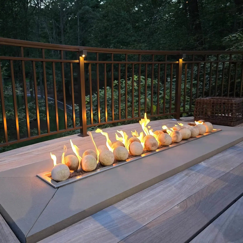 Linear gas fire feature on a deck at dusk, using Cottage White 4-inch ceramic fire balls with lively flames rising across the burner, framed by a wood railing and wooded backyard view.