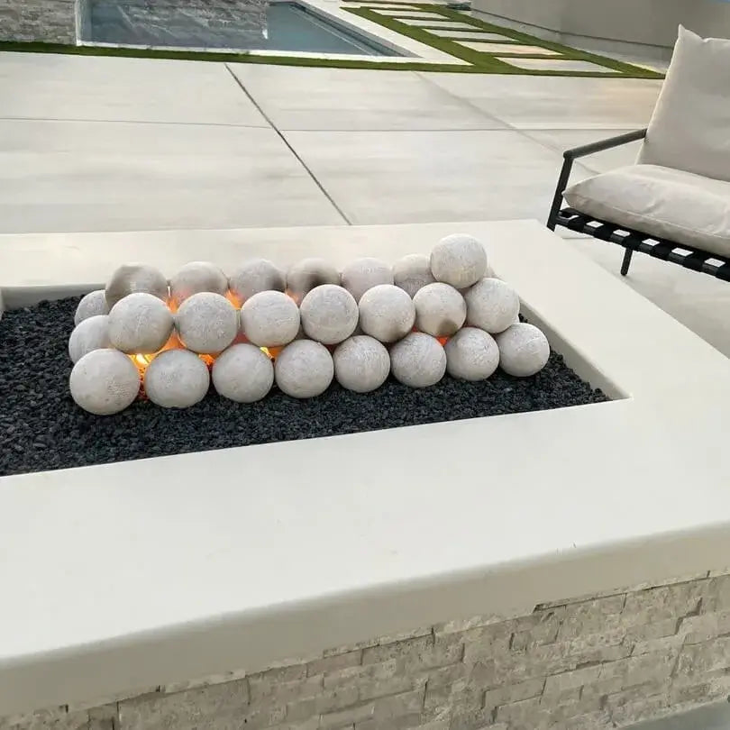 Modern rectangular concrete fire pit filled with black lava rock and stacked Cottage White 4-inch fire balls, with warm flames glowing between the stones beside a poolside patio chair.